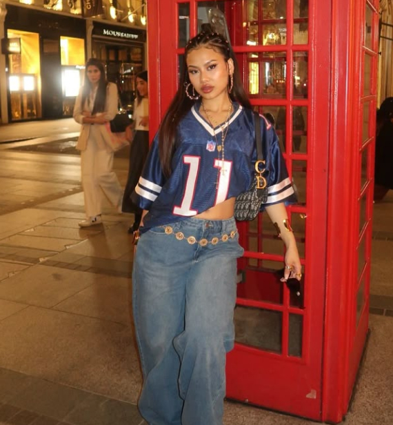 A person with braided hair poses next to a red telephone booth, wearing a cropped blue and white football jersey, wide-leg jeans with a chain belt, and layered necklaces, carrying a patterned handbag.