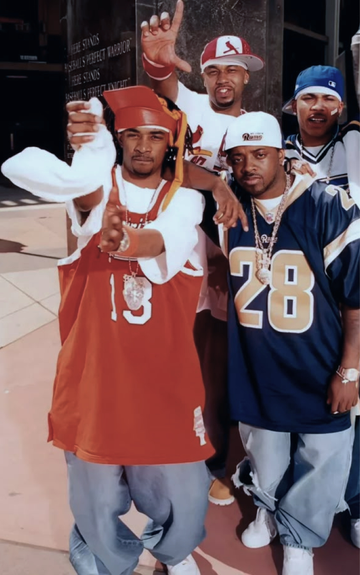 Four men, including Nelly and Murphy Lee, pose in 90s hip hop fashion, wearing oversized jerseys, baggy jeans, and baseball caps, flashing hand signs.