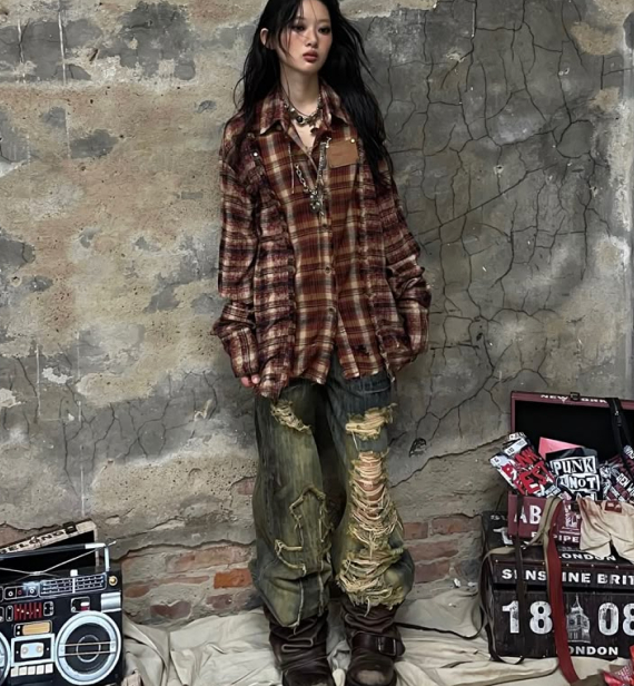 A person with long dark hair stands against a weathered wall, wearing an oversized plaid shirt, heavily distressed and ripped jeans, and chunky boots.