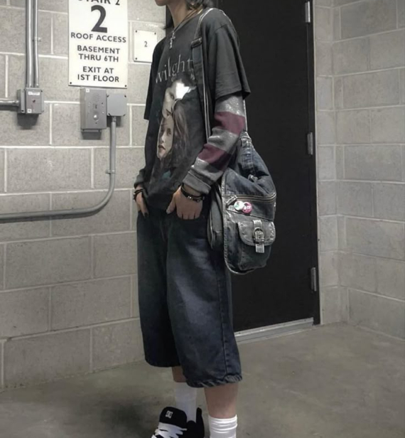 A person stands in a stairwell wearing a layered graphic t-shirt over a striped long-sleeve shirt, baggy denim shorts, sneakers, and a worn shoulder bag.
