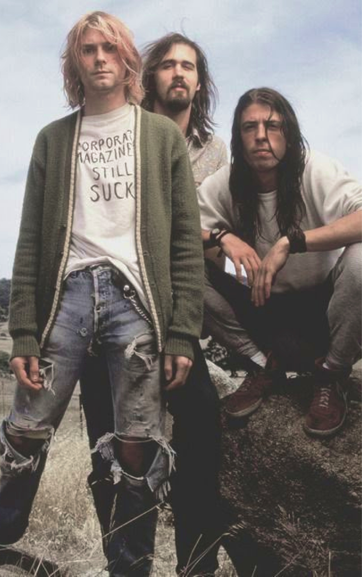 Nirvana, including Kurt Cobain, Krist Novoselic, and Dave Grohl, pose outdoors in grunge-era attire.