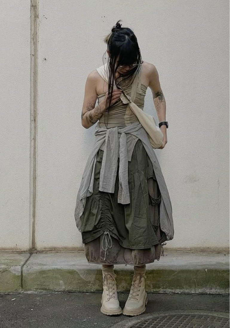 A person stands against a textured wall wearing a layered, neutral-toned outfit with a
                            draped top, olive green skirt with drawstrings, a tied gray garment, and off-white boots,
                            carrying a light-colored bag.